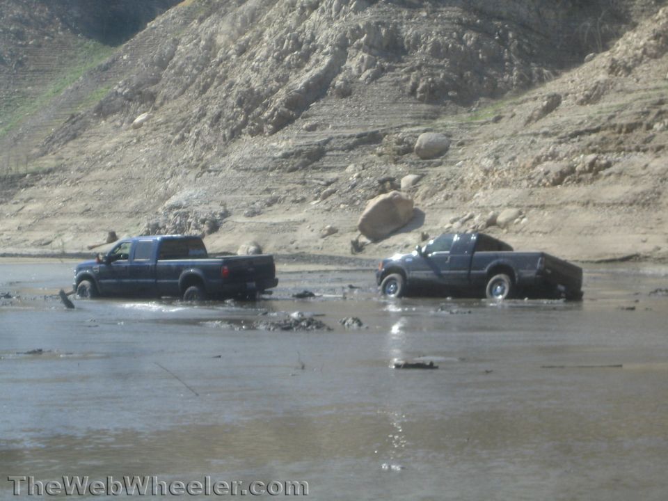 Two pickup trucks crossing a flooded muddy wash below a hillside