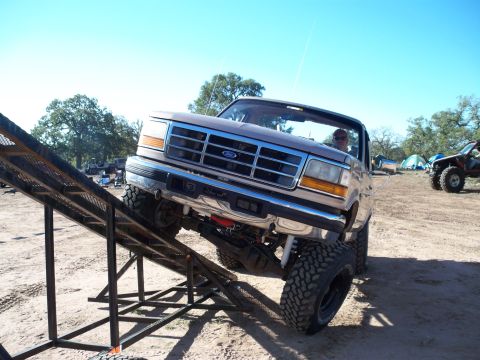 1996 Ford Bronco - A lifted, brown Ford Bronco is positioned on a metal ramp, with its front tires elevated. The vehicle is outdoors in a dirt lot with trees…