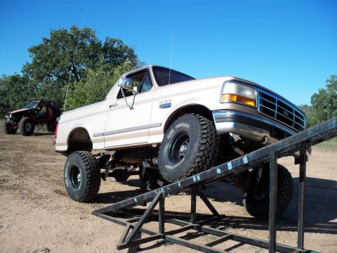 1996 Ford Bronco - A lifted, tan 1996 Ford Bronco is positioned on a metal ramp with its front tires elevated. Another off-road vehicle is visible in the back…