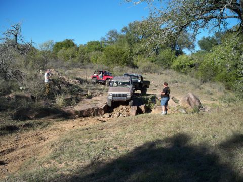 1996 Ford Bronco - A group of off-road vehicles navigate a rocky trail under a clear blue sky. A man in a black shirt and blue shorts stands near a tan Jeep C…