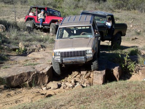 1996 Ford Bronco - A tan Jeep Cherokee is navigating a rocky obstacle, with its front tire on top of a large boulder. A red Jeep Wrangler and a dark green For…