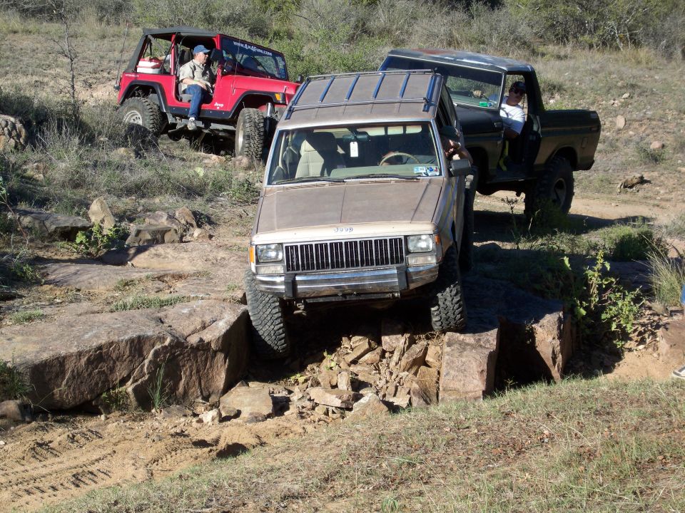 Tan Jeep Cherokee on rocks with red Jeep Wrangler and Ford Bronco behind.