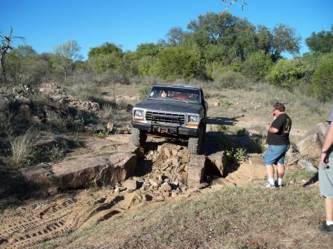 1996 Ford Bronco - A dark-colored Ford Bronco is navigating a rocky, off-road trail with a person standing nearby. The vehicle is positioned on large rocks, w…