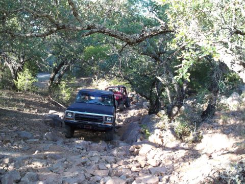 1996 Ford Bronco - Two off-road vehicles are navigating a rocky trail under a canopy of trees. The lead vehicle is a dark-colored SUV, possibly a Ford Bronco,…