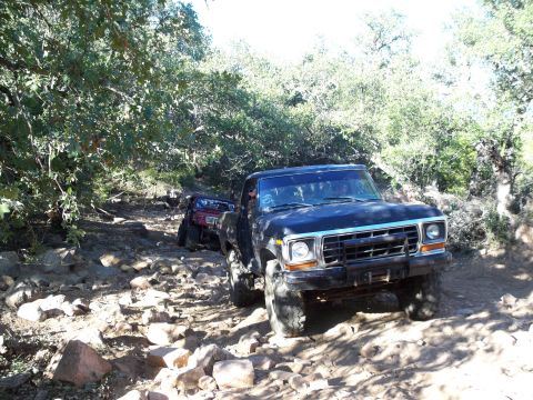 1996 Ford Bronco - A black Ford pickup truck is driving on a rocky, wooded trail. Another red off-road vehicle is visible further up the trail.