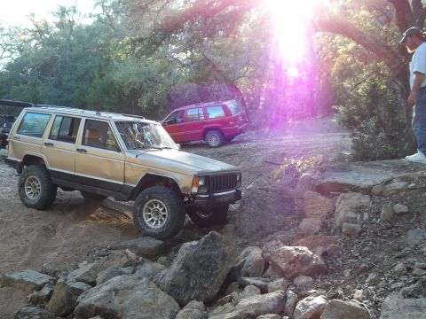 1996 Ford Bronco - A tan 1990s era Jeep Cherokee XJ is navigating a rocky, off-road trail with a red Jeep Cherokee XJ in the background. The sun is shining br…