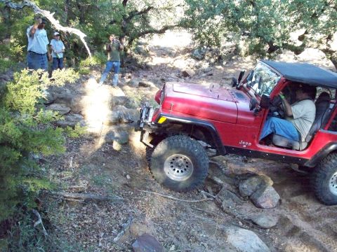 1996 Ford Bronco - A red Jeep Wrangler is navigating a rocky trail with several people standing nearby. The driver is visible inside the vehicle, gripping the…