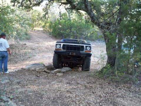 1996 Ford Bronco - A lifted, dark-colored Ford Bronco is navigating a rocky, off-road trail surrounded by trees. A person wearing a white t-shirt and blue jea…