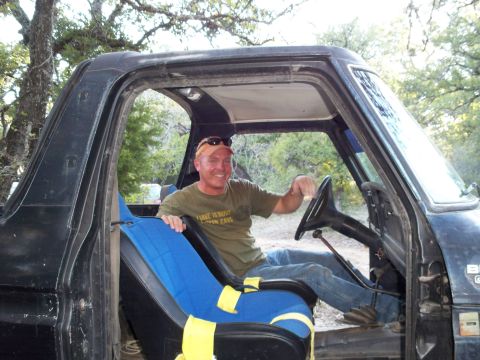 1996 Ford Bronco - A man wearing a tan baseball cap and sunglasses sits in the driver's seat of a black Ford Bronco, holding the steering wheel. The interior…