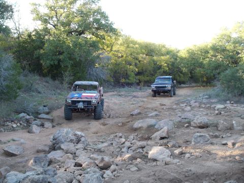 1996 Ford Bronco - Two trucks, including a 1996 Ford Bronco with Texas flag paint, navigate a challenging rocky trail during an off-road event.