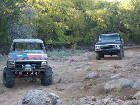 1996 Ford Bronco - Two lifted trucks, one painted with a Texas flag, navigate a rocky off-road trail.