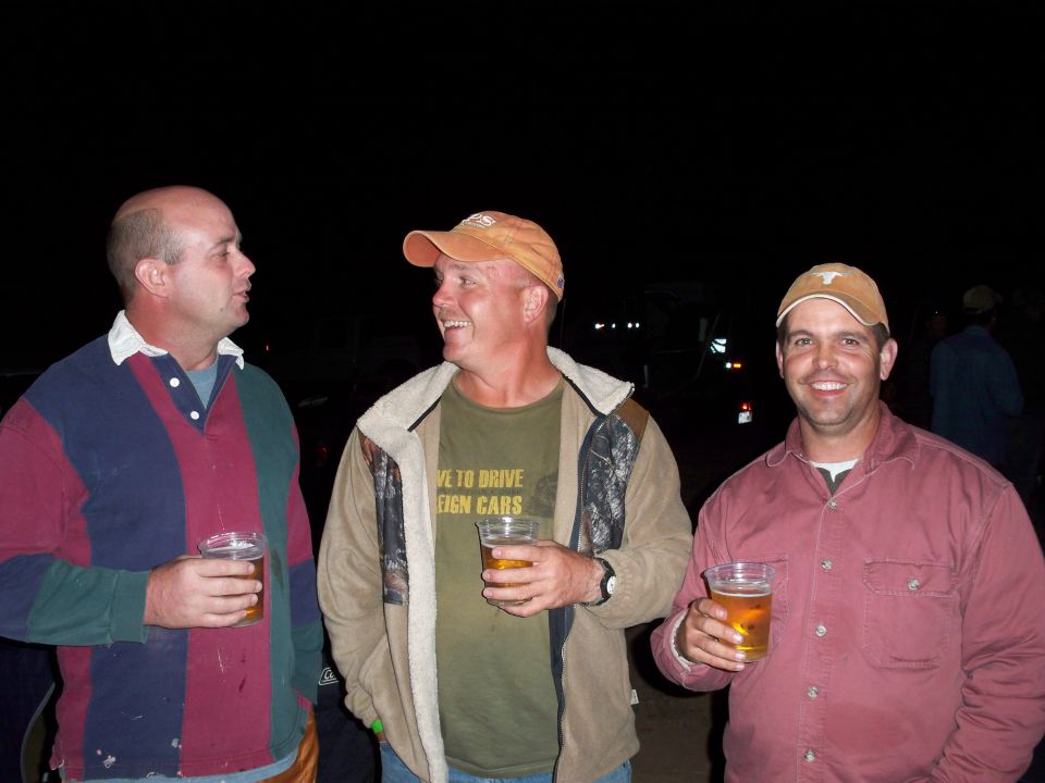 Three men holding beers at night, one wearing a shirt that says "I LOVE TO DRIVE FOREIGN CARS"