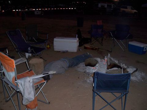 1996 Ford Bronco - A person is lying down near a campfire at dusk, surrounded by camping chairs and coolers.