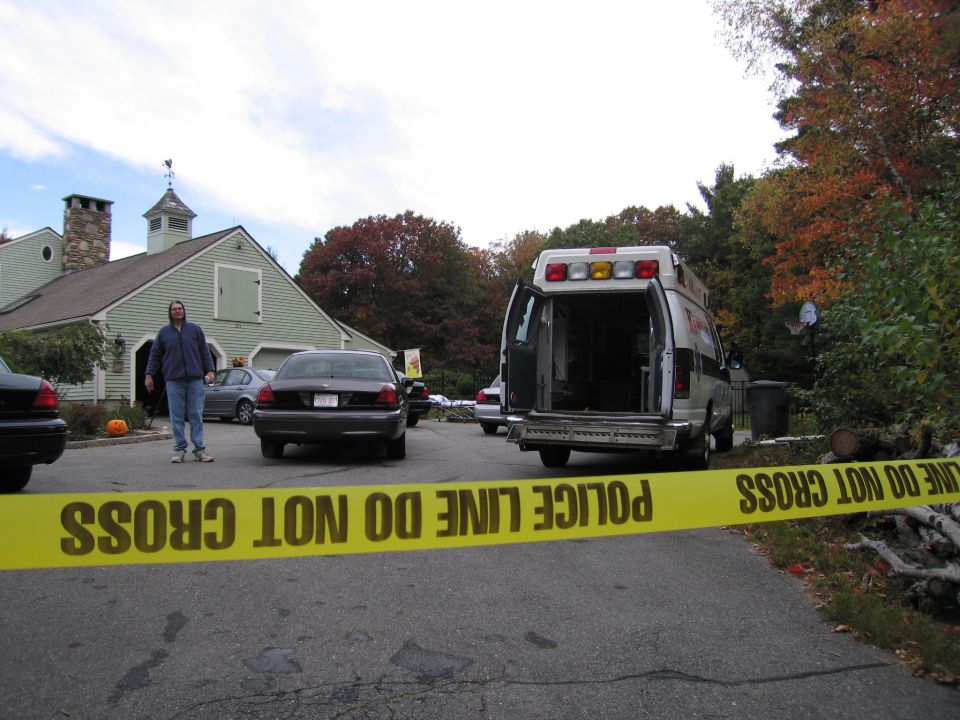 Police tape in foreground, two dark sedans and an ambulance in background. A man stands near a building