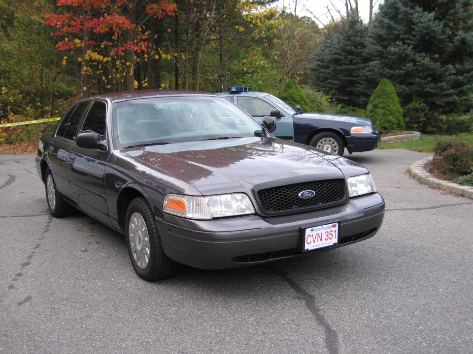 Dark gray 2001 Ford Crown Victoria, front 3/4 view, with police car behind