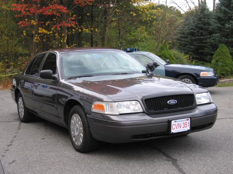 2001 Ford Crown Victoria - A dark gray 2001 Ford Crown Victoria police car is parked in front of another police car, with trees in the background.