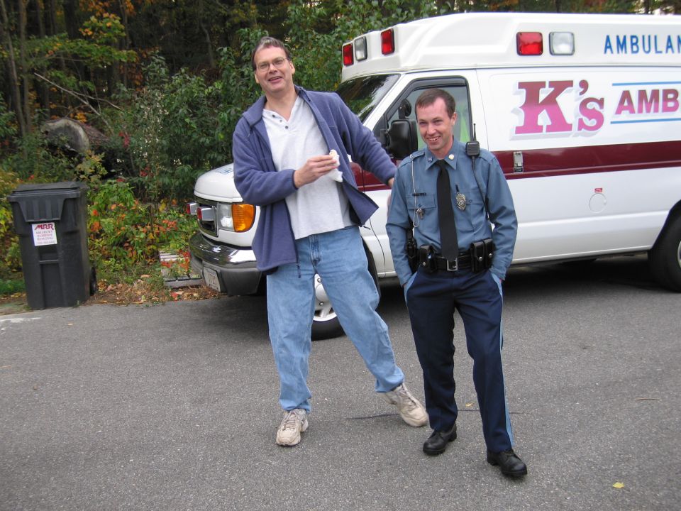 Man in blue jacket and jeans stands next to a police officer in front of an ambulance