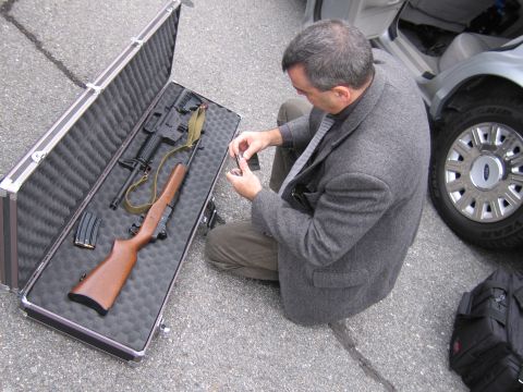 2001 Ford Crown Victoria - A man kneels beside an open gun case, preparing firearms next to a silver vehicle.