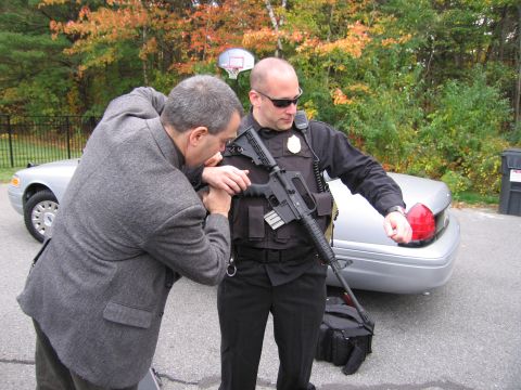 2001 Ford Crown Victoria - A civilian inspects a rifle carried by a police officer, with a silver sedan visible behind them.