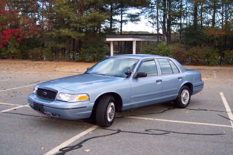 2001 Ford Crown Victoria - A light blue 2001 Ford Crown Victoria is parked in an empty lot with trees in the background.