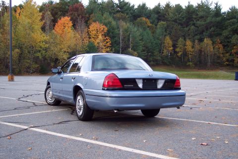 2001 Ford Crown Victoria - A light blue 2001 Ford Crown Victoria is parked in a lot during autumn, with colorful trees visible behind it.