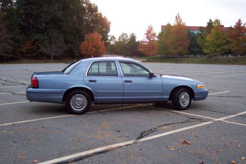2001 Ford Crown Victoria - A light blue sedan is parked in a lot during autumn, with trees showing fall colors.