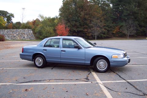 2001 Ford Crown Victoria - A blue 2001 Ford Crown Victoria sedan is parked in an outdoor lot with trees in the background.