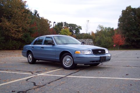 2001 Ford Crown Victoria - A blue 2001 Ford Crown Victoria sedan is parked in an empty lot with autumn foliage in the background.