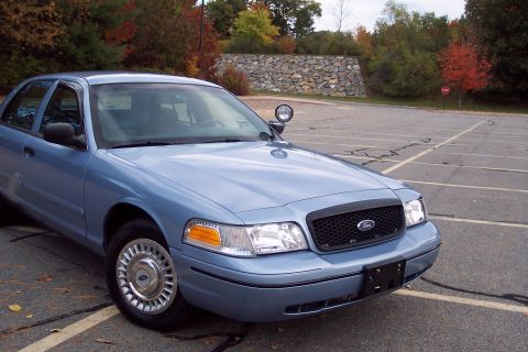 2001 Ford Crown Victoria - A blue 2001 Ford Crown Victoria is parked in a lot with autumn foliage in the background.