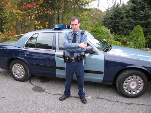 2001 Ford Crown Victoria - A police officer stands with arms crossed in front of a patrol car, with trees and foliage in the background.