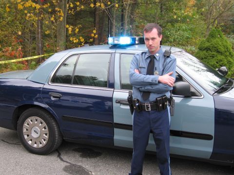 2001 Ford Crown Victoria - A police officer stands with arms crossed in front of a two-tone blue police car with its emergency lights on.