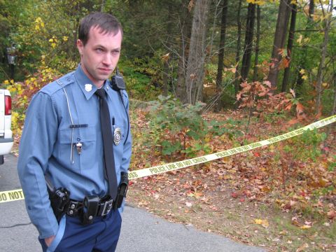 2001 Ford Crown Victoria - A police officer stands guard near caution tape in a wooded area, suggesting a crime scene investigation.