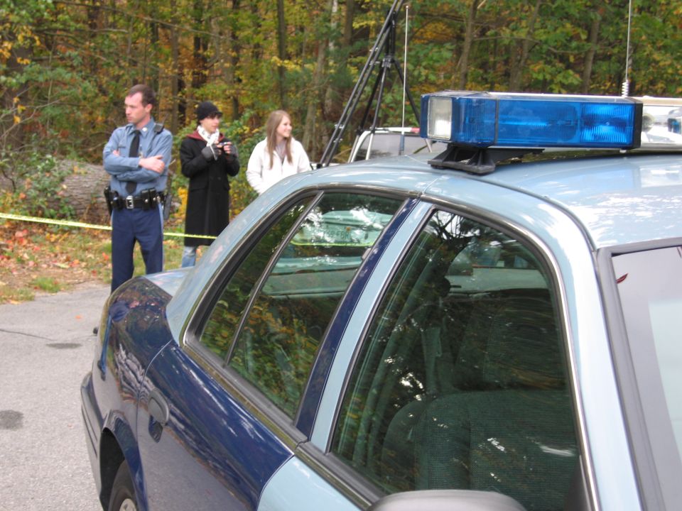 Blue and light blue police car side view, with blue light bar on roof. Officer and two civilians stand behind yellow tape
