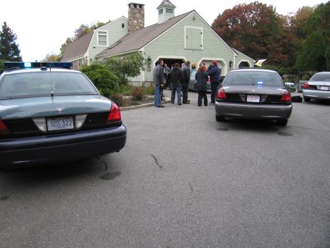 2001 Ford Crown Victoria - Two police cruisers are parked in a driveway in front of a large house, with several people gathered near the garage.