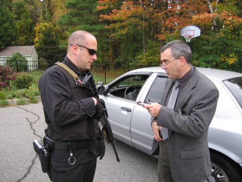 2001 Ford Crown Victoria - A police officer and a civilian converse outdoors next to a parked sedan.