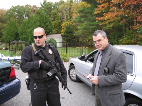 2001 Ford Crown Victoria - Two men, one in tactical gear with a rifle and another in a suit, stand beside a silver police car.