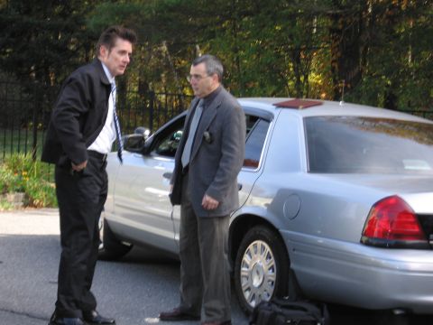 2001 Ford Crown Victoria - Two men in suits stand next to a silver 2001 Ford Crown Victoria sedan on a tree-lined street.