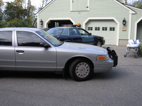 2001 Ford Crown Victoria - Two cars, one silver and one police-marked, are parked in a driveway next to a residential garage.