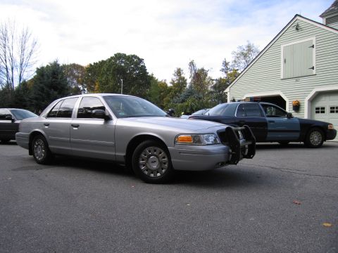 2001 Ford Crown Victoria - Two police cruisers, one silver and one two-tone, are parked in front of a residential garage.