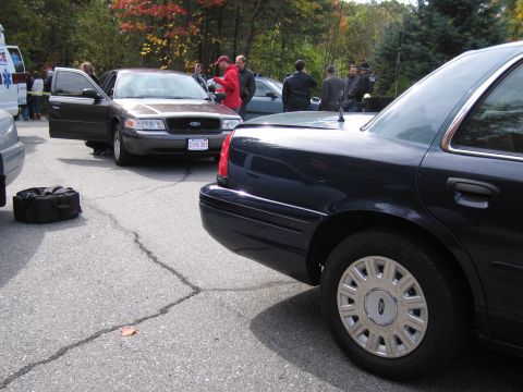 2001 Ford Crown Victoria - Two Ford Crown Victorias are parked in a lot, one with its door open and people gathered around.