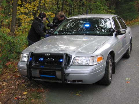 2001 Ford Crown Victoria - A silver police car is being filmed by a camera crew in a wooded area during the fall.