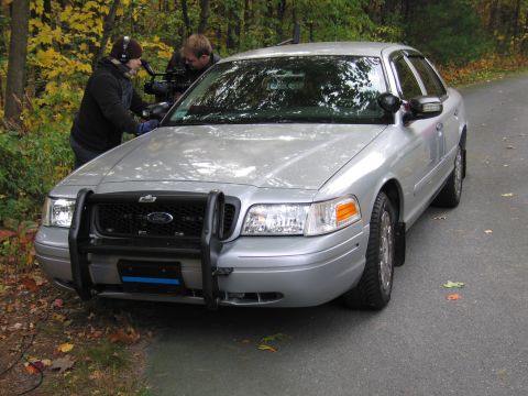 2001 Ford Crown Victoria - A silver 2001 Ford Crown Victoria is parked on a road while two people film it.
