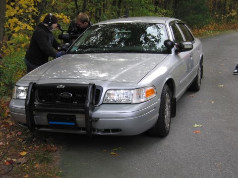 2001 Ford Crown Victoria - A silver 2001 Ford Crown Victoria is parked on a road, with a push bar on the front and two people filming in the background.
