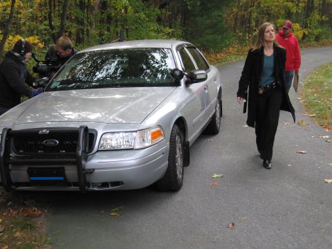 2001 Ford Crown Victoria - A silver Ford Crown Victoria police car is parked on a road with people filming.