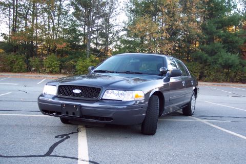 2001 Ford Crown Victoria - A dark gray 2001 Ford Crown Victoria is parked in an empty lot with trees in the background.