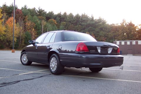 2001 Ford Crown Victoria - Rear view of a dark 2001 Ford Crown Victoria parked in an empty lot with trees in the background.