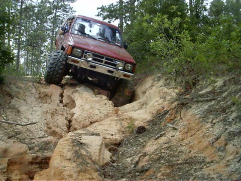 1985 Ford Bronco - A red SUV navigates a challenging dirt incline with large off-road tires.