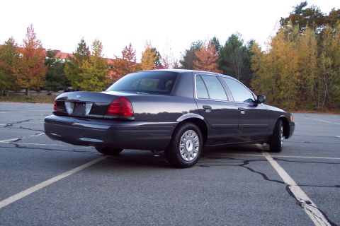 2001 Ford Crown Victoria - A dark gray sedan is parked in an empty lot with autumn foliage visible in the background.