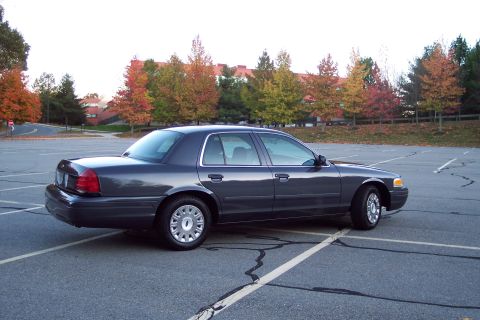 2001 Ford Crown Victoria - A dark gray 2001 Ford Crown Victoria sedan is parked in a lot with autumn trees in the background.