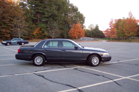 2001 Ford Crown Victoria - A dark gray 2001 Ford Crown Victoria sedan is parked in a lot, with another car visible in the background and autumn foliage.